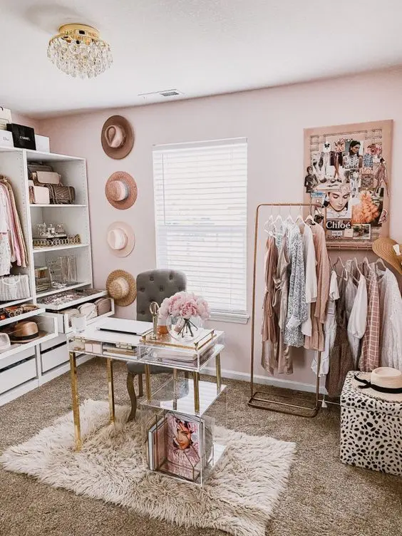 a lovely pink closet with open storage units and drawers, a makeshift clsoet, a crystal chandelier, a mini desk and a grey chair plus hats as decor