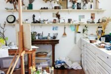 a messy and eclectic home art studio with open shelves with decor and dried foliage, a white storage cabinet, an easel, stools and console tables