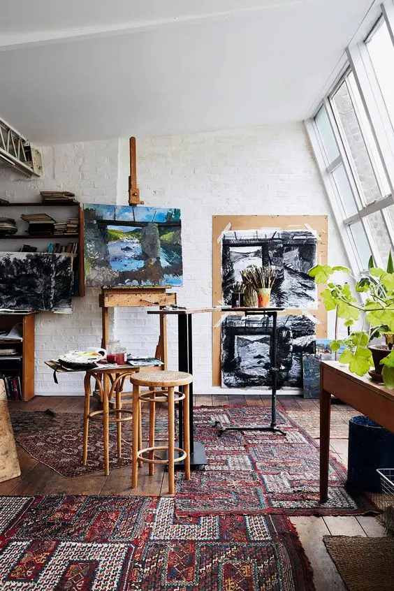 A mid century modern home artist studio with a glazed wall, colorful boho rugs, an easel, some stools, a large table and artwork