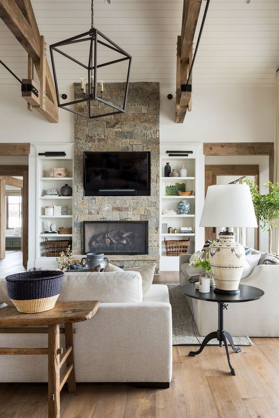 A neutral barn living room with a built in fireplace, neutral seating furniture, wooden beams, a metal chandelier and baskets