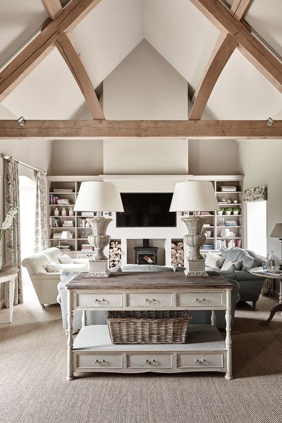 a neutral barn living room with a metal hearth, built-in shelves, neutral and pastel seating furniture, a whitewashed console table, wooden beams