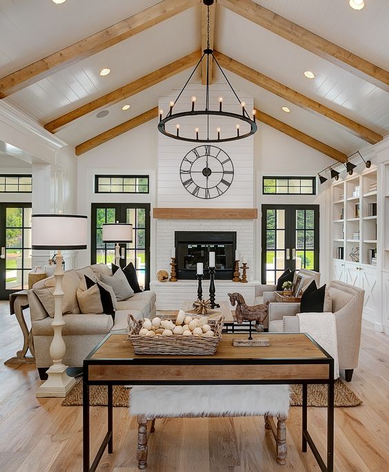 a neutral barn living room with white planked walls and a ceiling, wooden beams, a fireplace, neutral seating furniture, coffee tables and a metal chandelier