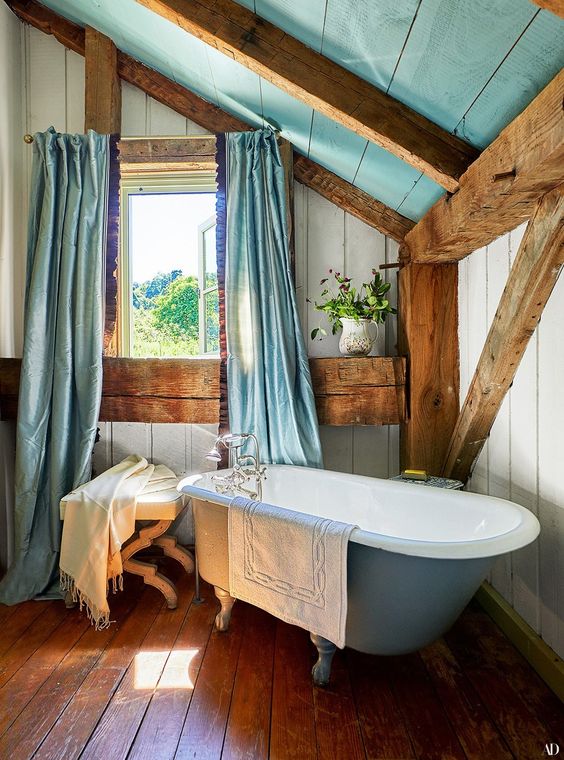 a pretty barn bathroom with wooden plank walls, a floor and ceiling, wooden beams, a blue clawfoot tub and blue curtains on the window