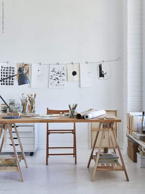 A pretty light filled studio with a trestle desk, a chair, a file cabinet, a table lamp, some shelves and lots of artwork