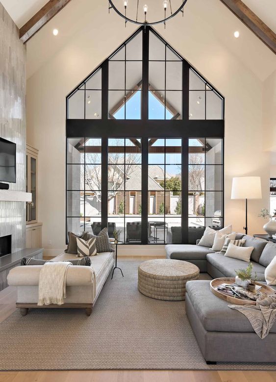 A refined modern living room with double height windows, a fireplace clad with tiles, a grey sectional, white daybed and a round pouf