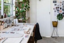 a simple and pretty art studio with a large table, a chair, potted plants, lots of art and art supplies is amazing