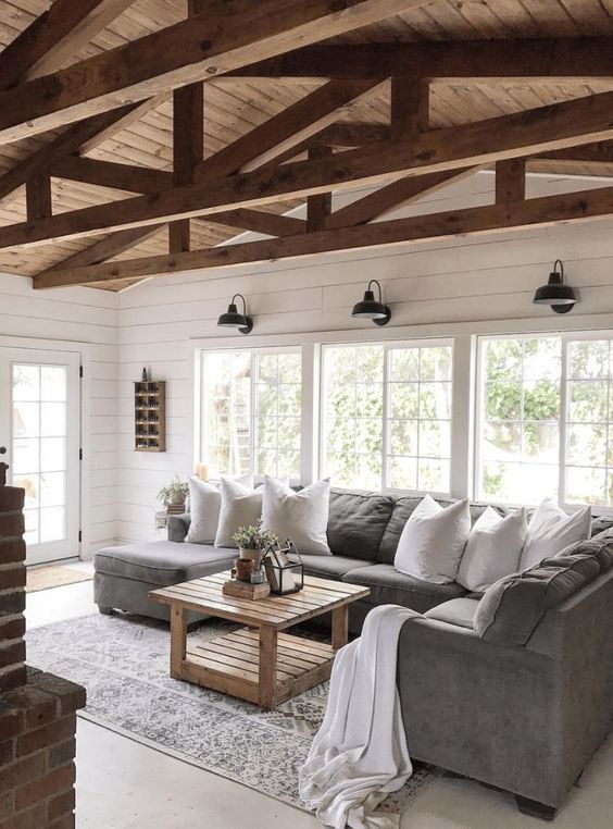 a simple barn living room with white planked walls, a grey sofa, wooden beams, a wooden table and lots of natural light