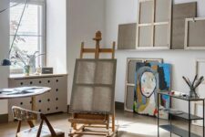 a simple home art studio with an easel, a chair, a table, a cart with supplies and some artwork – nothing unnecessary
