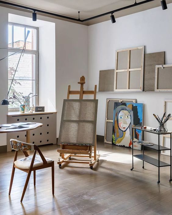 A simple home art studio with an easel, a chair, a table, a cart with supplies and some artwork   nothing unnecessary