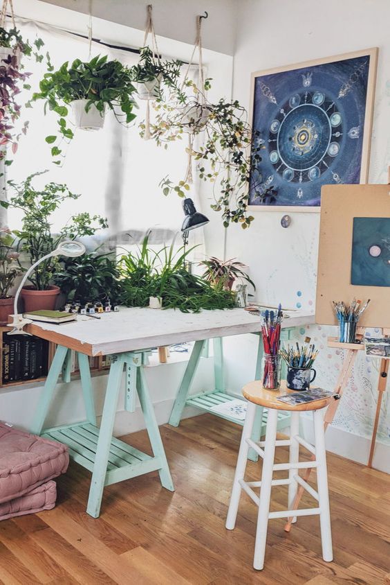 a small and cool art studio with a mint trestle desk, a stool, some cushions, an easel, potted plants and books