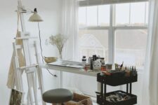 a small and cool home artist nook with a white console table, a black IKEA cart with supplies, a white easel, a white sconce and a stool