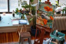 a small bright home artist studio with an easel, a trestle desk, a metal chair, a blue IKEA cart with supplies and lots of potted plants