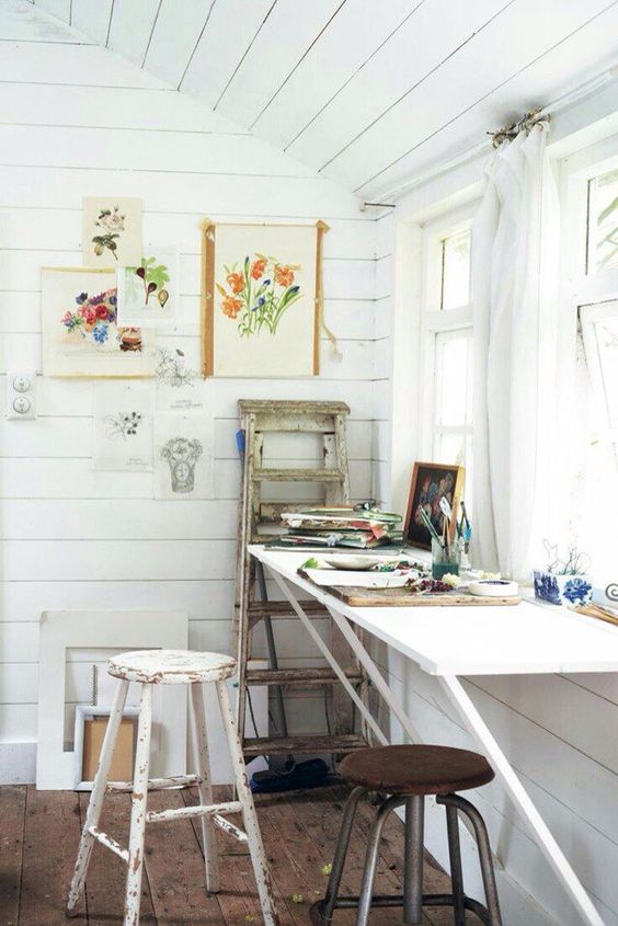 A small rustic art nook by the window with a wall mounted table, stools, a ladder, some art and art supplies