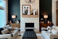 a sophisticated modern barn living room with a fireplace clad with faux stone, neutral seating furniture, a printed rug, a black ottoman, table lamps and a chic chandelier