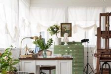 a vintage art studio with a white desk, vintage chairs and a footrest, a stained easel, a green file cabinet, potted plants and a crystal chandelier
