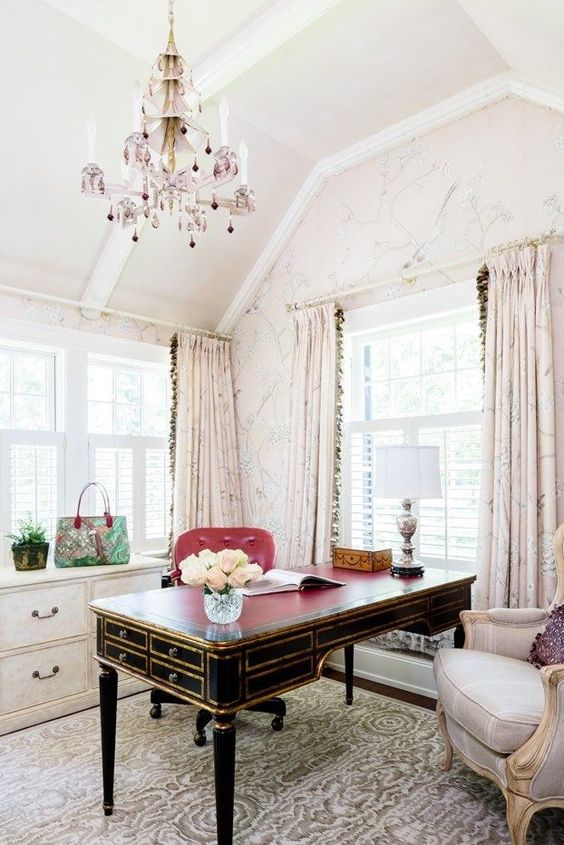 a welcoming and elegant feminine home office with blush floral walls and curtains, a refined black and gold desk, a lilac and pink chandelier and a chic chair