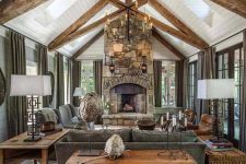 a welcoming barn living room with wooden beams, a fireplace clad with stone, grey seating furniture, a metal chandelier and baskets