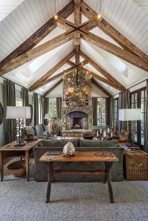 a welcoming barn living room with wooden beams, a fireplace clad with stone, grey seating furniture, a metal chandelier and baskets