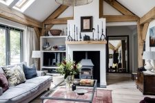 a welcoming barn living room with wooden beams,  a vintage hearth, grey seating furniture, a glass sofa, various accessories