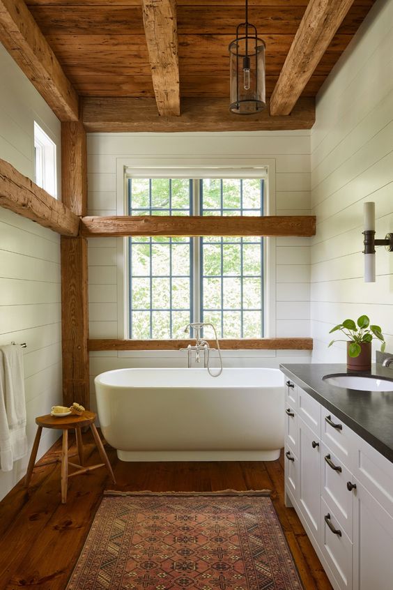 A white barn bathroom with wooden beams, a wooden ceiling with beams, a free standing tub, vintage furniture