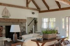 a white barn living room with a fireplace clad with stone, white seating furniture, dark stained tables, potted lavender in a basket and lots of wooden beams