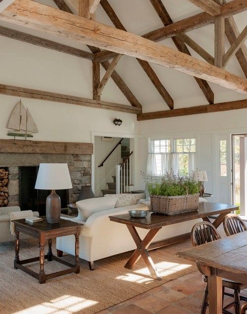 a white barn living room with a fireplace clad with stone, white seating furniture, dark stained tables, potted lavender in a basket and lots of wooden beams