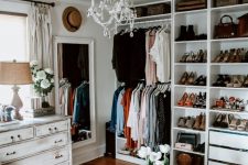 a white glam closet with open storage units and drawers, a shabby chic dresser, a large mirror, a crystal chandelier, a faux fur stool