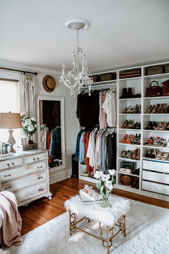 a white glam closet with open storage units and drawers, a shabby chic dresser, a large mirror, a crystal chandelier, a faux fur stool
