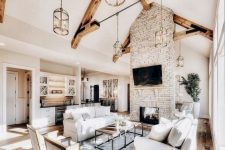 an airy barn living room with a brick fireplace, wooden beams, white seating furniture, a coffee table and lots of natural light incoming
