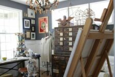 an art studio with a vintage stained file cabinet, a large metal desk, stools, a wooden easel, a chic chandelier