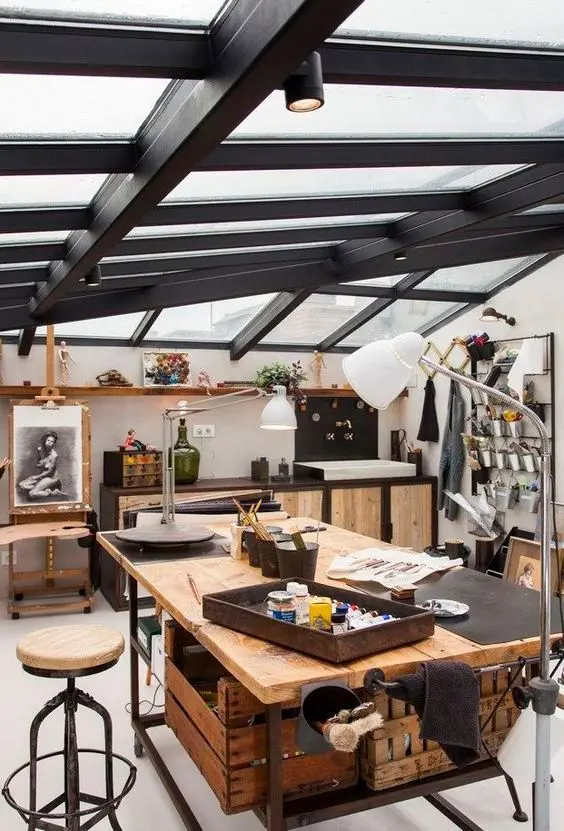 an industrial attic studio with a glazed roof, storage cabinets, a large working table and a matching stool, an easel and an organizer with metal buckets