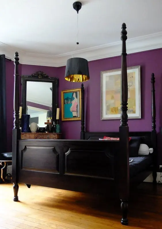 A bold purple bedroom with very dark furniture, a non working fireplace and a mirror, a pendant lamp and bold artworks