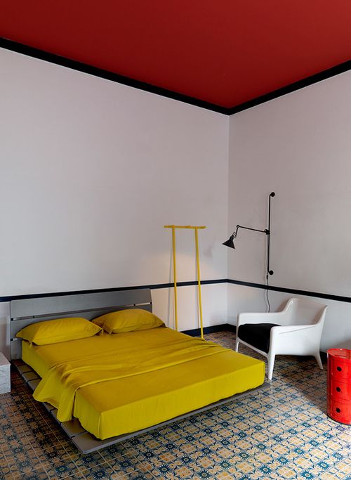 a catchy bedroom with a red ceiling, a grey bed with mustard bedding, a white chair, a red nightstand and a yellow stand
