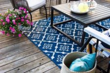 a colorful eclectic deck with a bright rug, wicker and wooden furniture, colorful textiles and bright blooms in planters