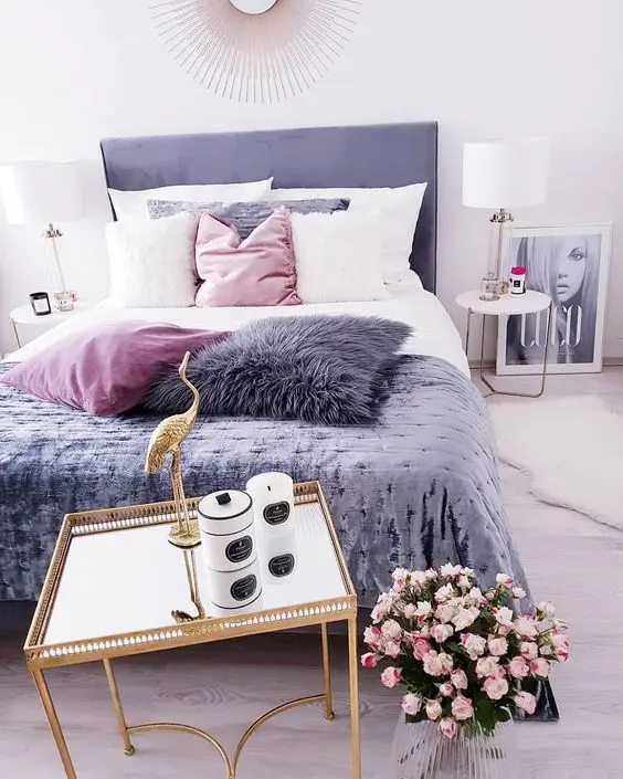 a contemporary glam bedroom in white, with a purple bed, pink and purple pillows, round nightstands and table lamp plus a mirror table