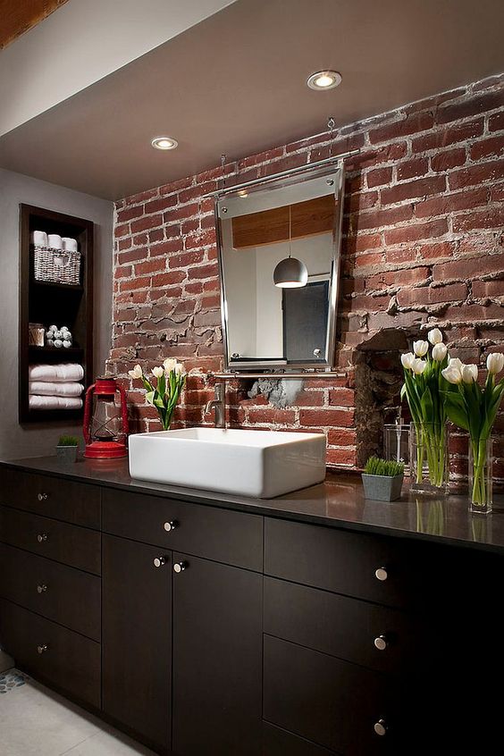 A dark bathroom with a red brick wall and a dark wooden vanity plus built in shelves