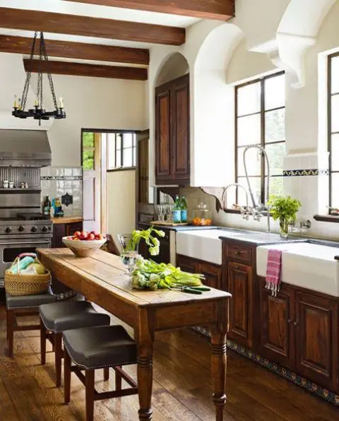 A dark stained shaker style kitchen with dark stone countertops, a ceiling with dark beams, a vintage stained table that doubles as a kitchen island