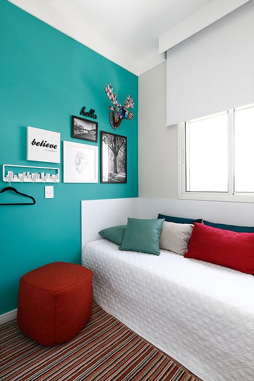 a lovely guest bedroom with a turquoise accent wall, a white daybed and bright pillows, a red pouf and a gallery wall