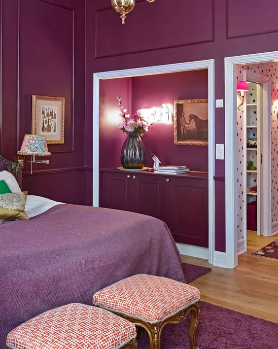 a luxurious purple bedroom done with paneling, a bed with purple and white bedding, refined stools and a niche for books