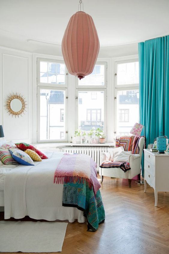 a neutral bedroom with a bay window, neutral furniture, colorful textiles, a pink pendant lamp and turquoise curtains