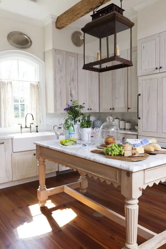 a pretty cottage whitewashed kitchen with white stone countertops, a neutral table that doubles as a kitchen island and a metal pendant lamp