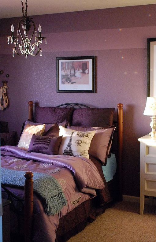 a purple bedroom with a forged bed, purple and pink bedding, a crystal chandelier and some artworks