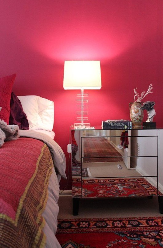 A refined mid century modern bedroom with a red accent wall, bold red and burgundy bedding, a printed rug and a mirror nightstand with a chic lamp