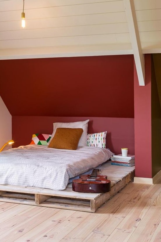 a small attic bedroom with a burgundy statement wall, a simple pallet bed, bright printed bedding and books