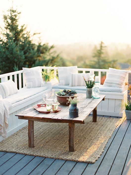 A small deck with a built in white wooden bench, a rustic coffee table and a jute rug is very welcoming to receive guests