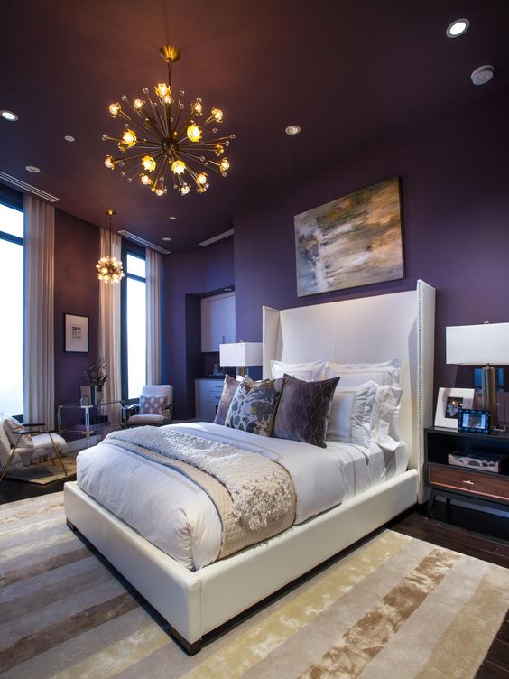 a sophisticated bedroom with deep purple walls, a white bed, stained furniture, sunburst chandeliers and some artworks