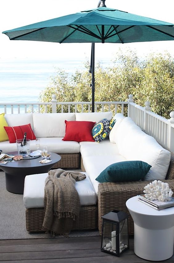A welcoming summer deck with an L shaped wicker sofa, a coffee table, some lanterns and a teal umbrella
