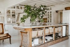 a white cottage kitchen with glass cabinets, a large antique kitchen island with plenty of storage space and greenery