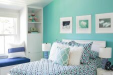 an attic bedroom with a turquoise accent wall, a white bed and colorful bedding, a windowsill bench and built-in storage units