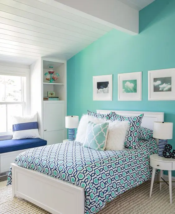 An attic bedroom with a turquoise accent wall, a white bed and colorful bedding, a windowsill bench and built in storage units
